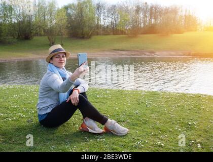 Frau in grauem Oberteil, Strohhut, schwarze Hose/Hose, blauer Schal, der am Telefon fotografiert und lächelt, auf Wiese/grünem Gras am Wasser sitzend Stockfoto
