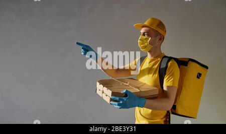 Sichere Lieferung von Lebensmitteln. Kurier in gelber Uniform, Schutzmaske und Handschuhe liefert während der Coronovirus-Quarantäne Essen zum Mitnehmen. Nach links zeigend. Stockfoto