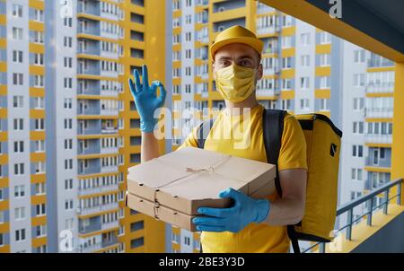 Sichere Lieferung von Lebensmitteln. Kurier in gelber Uniform, Schutzmaske und Handschuhe liefert während der Coronovirus-Quarantäne Essen zum Mitnehmen Stockfoto