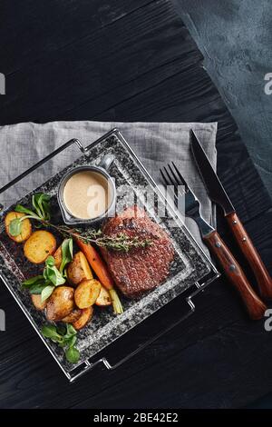 Fleischsteak mit gegrilltem Gemüse auf einer Marmorplatte. Saftiges Fleischsteak auf einem wärmenden Teller, Holzhintergrund, Draufsicht, mit Steakgeräten Stockfoto