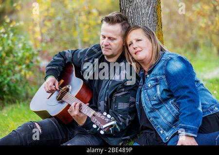 Ein reifes Paar verbringt viel Zeit zusammen und die Frau hört ihrem Mann singen und spielen seine Gitarre, während in einem Stadtpark auf einem ... Stockfoto