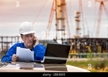 Mann arbeitet am Laptop und Smartphone, während er an einem Fahrzeug mit einer Ölplattform im Hintergrund steht; Alberta, Kanada Stockfoto