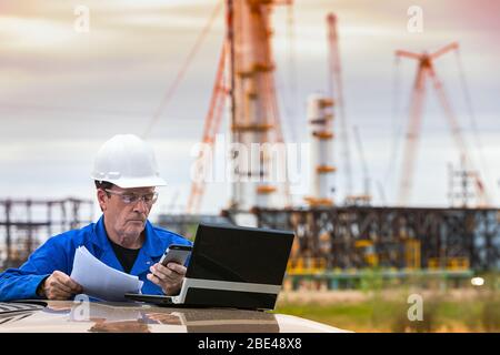 Mann arbeitet am Laptop und Smartphone, während er an einem Fahrzeug mit einer Ölplattform im Hintergrund steht; Alberta, Kanada Stockfoto