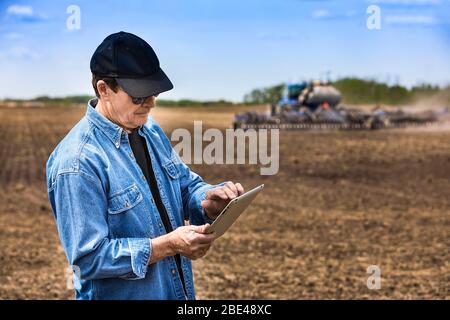 Landwirt mit einer Tablette, während auf einem Feld Bauernhof stehen und ein Traktor und Ausrüstung Samen das Feld; Alberta, Kanada Stockfoto