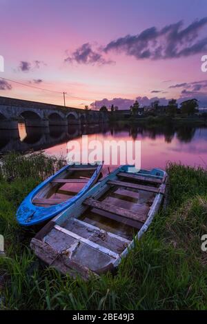 Zwei alte Holzboote am Ufer des Shannon River bei Sonnenuntergang mit einer Steinbrücke im Hintergrund; Montpellier, County Limerick, Irland Stockfoto