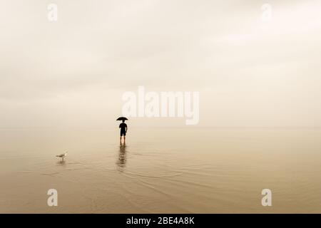 Mann, der am Strand mit einem Regenschirm im Regen steht. Kopieren Sie den Bereich für Inspiration Zitat oder Text Stockfoto