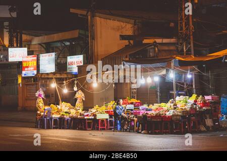 Eine ältere Verkäuferin sitzt mit ihrem Handy auf einem Nachtmarkt; Ho Chi Minh City, Vietnam Stockfoto