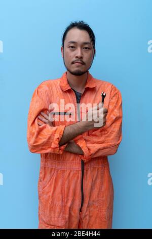 Der Chefmechaniker in orangefarbener Uniform hielt einen kleinen Schraubenschlüssel. Mit gekreuzten Armen auf dem blauen Bildschirm stehen. Porträt mit Studiolicht. Stockfoto