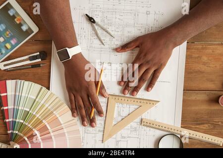 Architekt arbeitet an einem Entwurf. Draufsicht auf afro amerikanischen Mann, der etwas auf einem Blaupause während sitzen an seinem Arbeitsplatz zu Hause zeichnen. Skizzenplan wird gezeichnet. Freiberuflich. Arbeitsplatz im Home Office Stockfoto