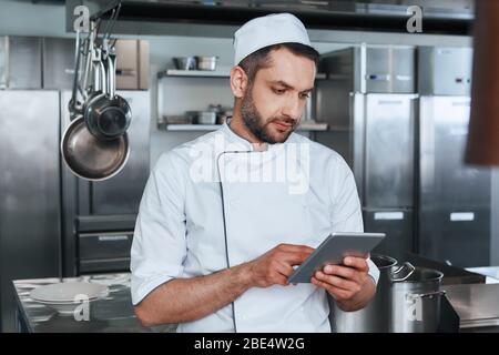 Bärtiger männlicher Koch schaut sich digitale Tablette an, während er Essen in der Restaurantküche zubereitet Stockfoto