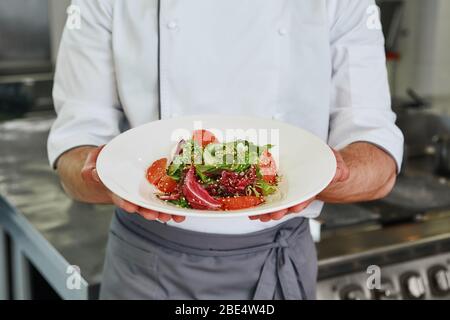 Der kaukasische Koch hat im Restaurant Salat zubereitet. Er steht in der kommerziellen Küche und hält sein Gericht. Zugeschnittenes Bild. Vorderansicht Stockfoto