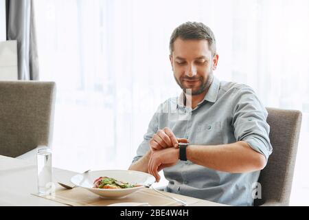 Bärtiger kaukasischer Mann wird Salat in seinem Büro essen. Er schaut auf seine Uhr, während er bei der Arbeit zu Mittag essen kann. Stockfoto