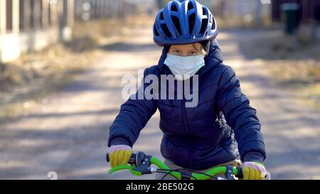 Ein Kind auf einem Spaziergang während der Quarantänezeit des Coronavirus Covid19. Der Junge fährt ein Fahrrad in einem hellblauen Helm. Kind tragen medizinische Schutzmaske und Handschuhe. Sonniger Tag. Stockfoto