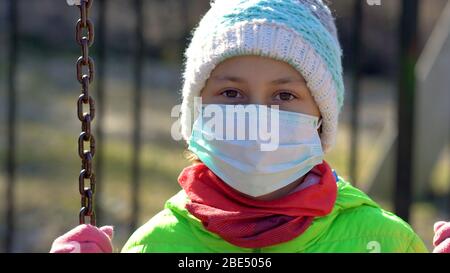 Mädchen auf einem Spaziergang während der Quarantänezeit des Coronavirus. Das Kind schwingt auf einer Kettenschaukel in einer medizinischen Schutzmaske und Handschuhen. Heller sonniger Tag. Stockfoto