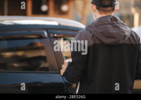 Kurier in Schutzmaske und medizinische Handschuhe nehmen Bestellung aus dem Auto. Lieferung Junge halten Pizza und Kaffee. Lieferservice unter Quarantäne. Coronavirus Stockfoto