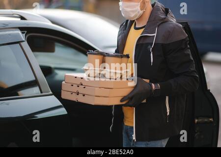 Kurier in Schutzmaske und medizinische Handschuhe nehmen Bestellung aus dem Auto. Lieferung Junge halten Pizza und Kaffee. Lieferservice unter Quarantäne. Coronavirus Stockfoto