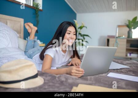 Junge Frau im Bett liegend mit gekreuzten Beinen, auf Laptop tippen, lächelnd Stockfoto