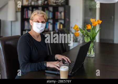 Kaukasierin mittleren Alters arbeitet von zu Hause aus auf einem Laptop in ihrem Wohnzimmer, trägt Gesichtsschutzmaske, während in Quarantäne und Isolation Stockfoto