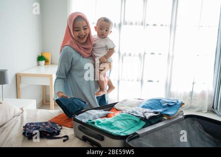 Muslimische asiatische Mütter tragen ihre Kinder, während sie Kleidung vorbereiten, um sie vor der Reise in ihre Koffer zu legen Stockfoto