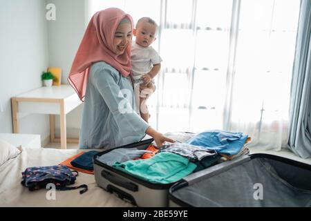 Verschleierte Mütter tragen ihre Kinder, während sie Kleidung im Schlafzimmer vorbereiten, bevor sie zur Reise gehen Stockfoto