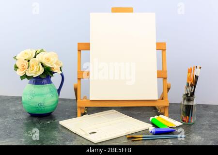 EASTRIGGS, SCHOTTLAND - 7. Januar 2020: Desktop-Easel und Leinwand, Pinsel, Acrylfarben und Vase mit weißen Rosen Stockfoto