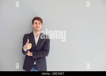 Lächelnder Mann Daumen nach oben in einem lässigen Jacke positive fröhlich fröhlich fröhlich fröhlich fröhlich auf einem grauen Copyspace Hintergrund. Stockfoto