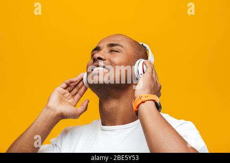 Porträt eines attraktiven lächelnden selbstbewussten jungen afrikanischen Mannes, der auf gelbem Hintergrund steht und Musik mit kabellosen Kopfhörern hört Stockfoto