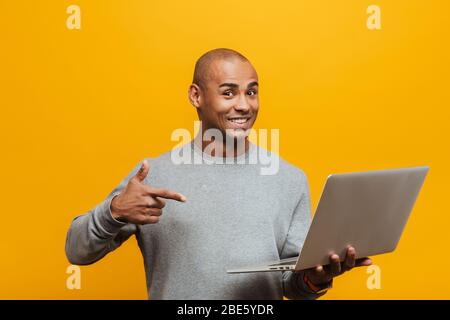 Porträt eines attraktiven lächelnden zuversichtlich lässig jungen afrikanischen Mann steht über gelben Hintergrund, zeigt auf Laptop-Computer Stockfoto