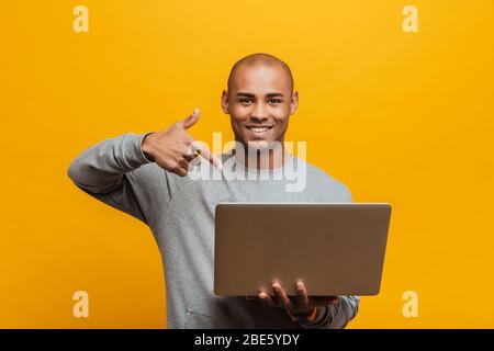 Porträt eines attraktiven lächelnden zuversichtlich lässig jungen afrikanischen Mann steht über gelben Hintergrund, zeigt auf Laptop-Computer Stockfoto