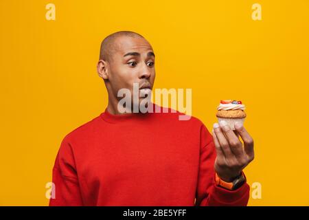 Porträt eines attraktiven schockierten lässig jungen afrikanischen Mann steht über gelben Hintergrund, Blick auf Cupcake Stockfoto