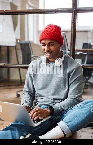 Bild eines positiv optimistischen jungen afrikanischen Mannes, der mit Laptop und Handy drinnen auf dem Boden sitzt. Stockfoto