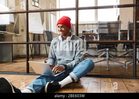Bild eines positiv optimistischen jungen afrikanischen Mannes, der mit Laptop und Handy drinnen auf dem Boden sitzt. Stockfoto