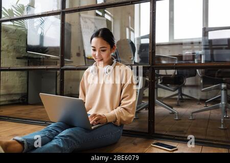 Bild von jungen schönen asiatischen Frau auf Laptop arbeiten, während auf dem Boden im Büro sitzen