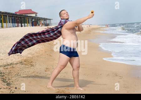 Am Strand steht ein witziger fetter Mann-Superheld mit Plaid statt Mantel und Bun in der Hand. Stockfoto