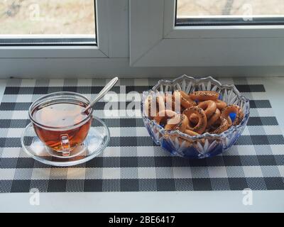 Auf der Fensterbank befindet sich eine transparente Tasse Tee und eine Schüssel Bagels. Unterhaltung in Isolation. Stillleben. Stockfoto