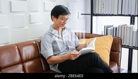Senior Asian Mann Lesen Buch auf Sofa im Wohnzimmer zu Hause, Porträt von asiatischen älteren Mann ist entspannend und Glück mit Lesen Sie ein Magazin zu Hause Stockfoto