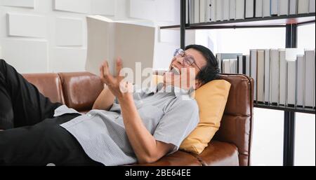 Älterer Asienmann liest Buch und lacht auf Sofa im Wohnzimmer zu Hause Porträt des asiatischen Älteren Mann ist entspannend und glücklich mit Read a Magazin Stockfoto