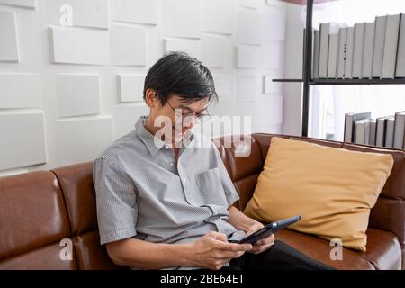 Senior Asian man spielt Tablet und Video Call auf dem Sofa im Wohnzimmer zu Hause Portrait des asiatischen älteren Mannes ist Entspannend und Glücklich Mit dem Spielen von TA Stockfoto