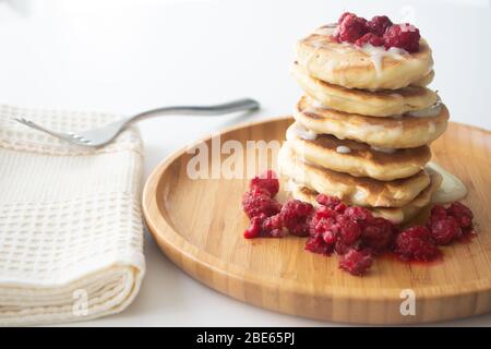 Flauschige Pfannkuchen mit Kondensmilch und Himbeeren auf einem Holzteller mit Küchenteller und Gabel Stockfoto