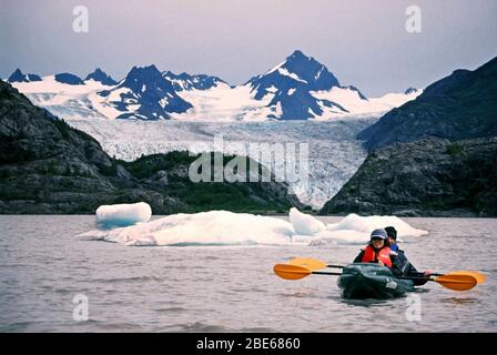 Mutter und Sohn in warmen Jacken, Hüten und Rettungswesten paddeln an einem Sommertag mit dem Kajak an Eisbergen in Halibut Cove, Homer, Alaska, USA vorbei. Stockfoto