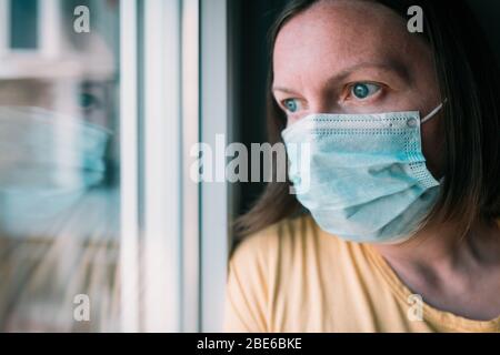 Frau in Selbstisolation während des Virenausbruchs, die durch das Fenster schaut. Besorgte weibliche Person mit schützenden chirurgischen Maske in zu Hause bleiben Konzept, sel Stockfoto