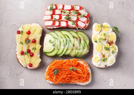 Fünf Sandwiches auf Toast mit frischen Karotten, Gurken, Ananas, rotem Currant, Krabbenstäbchen und Wachteleiern mit Erbsen Mikrogrün auf grauem Hintergrund Stockfoto