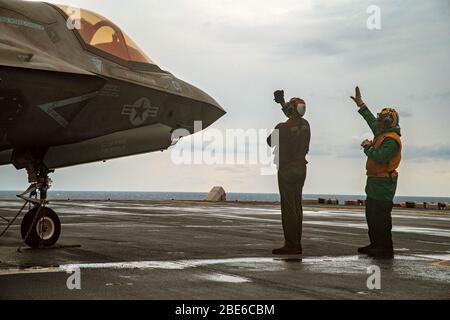 Die Matrosen der US-Marine geben dem Piloten eines Marine Corps F-35B Lightning II Stealth-Kämpfers, der der 31. Marine Expeditionary Unit zugewiesen wurde, Handsignale, nachdem eine vertikale Landung auf dem Flugdeck des amphibischen Amphibienschiffs USS America 9. April 2020 in der philippinischen See stattfand. Stockfoto