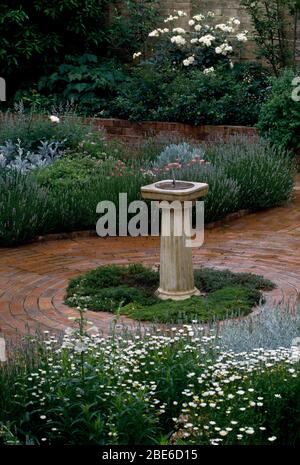 Steinsonnenuhr in der Mitte des gepflasterten Innenhof mit weißen Gänseblümchen und Rosen in Betten mit Lavendel eingefasst Stockfoto