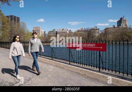 New York, NY, USA. 11. April 2020. Ein Paar in Schutzmasken geht an einem sozialen Distanzzeichen auf der Laufstrecke am Stausee in Central Par vorbei Stockfoto