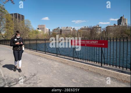 New York, NY, USA. 11. April 2020. Eine Frau in einer Schutzmaske geht an einem sozialen Distanzzeichen auf der Laufstrecke am Stausee in Central Par vorbei Stockfoto