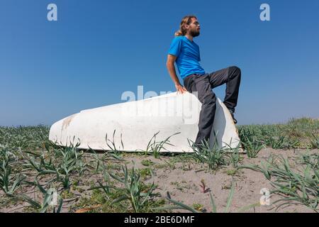 Ein Mann sitzt auf einem weißen Boot auf dem Sand. Es gibt Pflanzen im Sand. Der Himmel ist klar und blau. Stockfoto