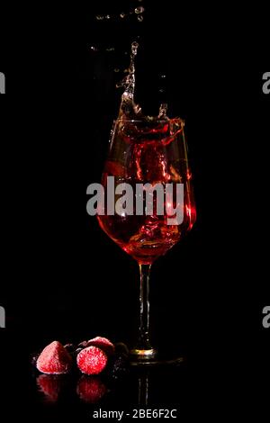 Rot- oder Roséwein, der aus einem Weinglas vor schwarzem Hintergrund mit Beeren spritzt. Weinspritzer, leise Stockfoto