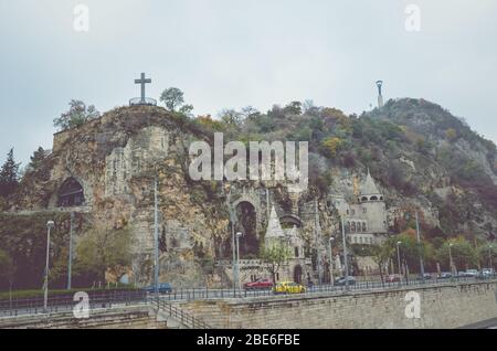 Budapest, Ungarn - 6. November 2019: Gellert Hill Cave Church, Sziklatemplom. Freiheitsstatue auf dem Hügel. Autos auf der Straße vor dem Hügel. Horizontales Foto. Ungarische Sehenswürdigkeiten. Stockfoto