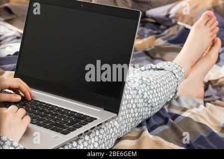 Vorderansicht auf einem Bildschirm eines Laptops auf den Knien einer Frau in einer grau blühenden Nachtwäsche. Home Office-Konzept. Freelancer arbeitet während in einem Bett Stockfoto